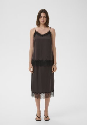 Kobieta w czarnej satynowej koszulce na cienkich ramiączkach i dopasowanej spódnicy o długości midi z koronkowym wykończeniem, stojąca boso na jasnym, jednolitym tle.