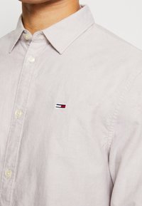 Lichtbeige katoenen overhemd met knoopsluiting en een omgeslagen kraag, met een klein geborduurd logo in rood, wit en blauw.