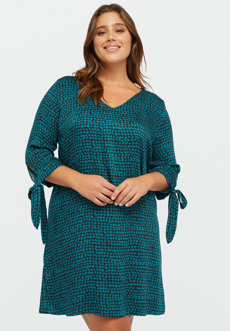 Robe turquoise avec un décolleté en V, des manches trois-quarts nouées aux poignets, et un motif à pois sur un fond sombre. Texture de tissu douce.