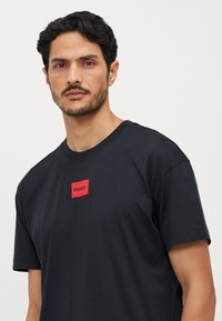 T-shirt en coton noir avec une étiquette rectangulaire rouge portant le texte "HUGO". Design à col ras du cou et manches courtes. Texture lisse, coupe décontractée.