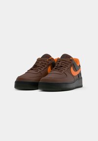 Smeđe kožne tenisice s narančastim naglascima, crnim potplatom, teksturiranim gornjim dijelom, okruglim vrhom i tradicionalnim vezanjem. Karakteristični Nike logo.
