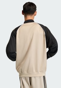 Beež sportlik jakk mustade võrgusvarrukatega, millel on valged aktsendivoodrid ja mugav lõige. Tekstuur on sile ning disain sportlik.