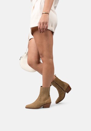 Bottines en suède marron avec une finition texturée, bout pointu, talon empilé et détail de zip sur le côté. Portées avec un short de couleur claire.