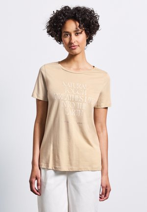T-shirt en coton beige à manches courtes et col rond. Présente un texte blanc qui dit "UN TOUCHER NATUREL INSPIRE LA VIE SUR TERRE."