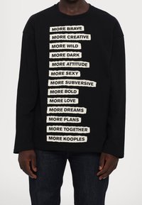 Felpa nera a maniche lunghe con testo sovrapposto color crema in grassetto e in stile ritagliato che esprime diverse affermazioni allineate verticalmente.