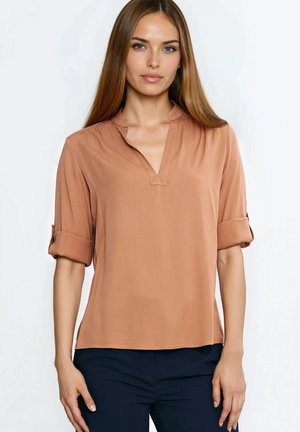 Młoda kobieta z długimi prostymi włosami, ubraną w brązową bluzkę z podwiniętymi rękawami i ciemne spodnie, stojąca na tle jednolitego jasnego tła.