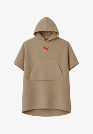 Mann trägt einen khakifarbenen Kapuzen-Handtuchponcho mit rotem Puma-Logo, Hände in der Vordertasche, steht vor einem einfarbigen Hintergrund.