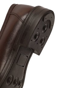 Brown leather shoe with a textured upper, curved shape, and a thick black rubber sole featuring circular indentations.