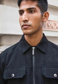 Schwarze Wildlederjacke mit Reißverschluss, spitzem Kragen, zweiFronttaschen und silbernen Hardware-Details. Glatte Textur und moderner Schnitt.