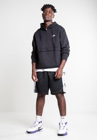 Zwarte pullover hoodie met een half-rits en een voorkantzak, gecombineerd met zwarte mesh shorts. Witte sneakers met paarse accenten maken de look af.