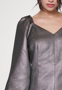 Metallinhohtoinen harmaa mekko, jossa on teksturoitu pinta, V-aukko ja liehuvaolkaimet olkapäällä, näyttäen monimutkaisia geometrisia kuvioita.