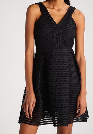 Femme portant une robe noire texturée sans manches, avec un décolleté en V et une jupe évasée, debout sur un fond uni.