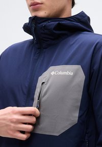 Άτομο που φοράει μπουφάν ναυτικού μπλε χρώματος με γκρι τσέπη στο στήθος με φερμουάρ και ετικέτα Columbia, κρατώντας το φερμουάρ της τσέπης σε κοντινή λήψη.