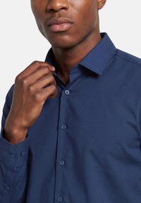 Mees kohendab nööbitud tumedat sinist kleidisärki, näidatud kaelast keskmise kehapiirini tavalise taustaga.