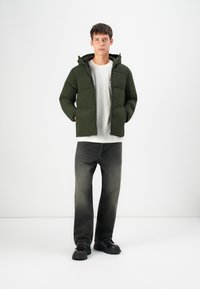 Puffer jacket in dark green, featuring a hood, zip closure, and front pockets. Paired with faded gray jeans and black chunky shoes.