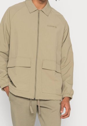 Beige lightweight jacket with a pointed collar, front zipper, and two large patch pockets. Elastic cuffs and minimal branding on chest.