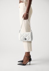 Borsa a tracolla bianca trapuntata con logo argentato, dotata di una maniglia corta, sostenuta di lato. Il modello indossa pantaloni chiari e tacchi alti neri.