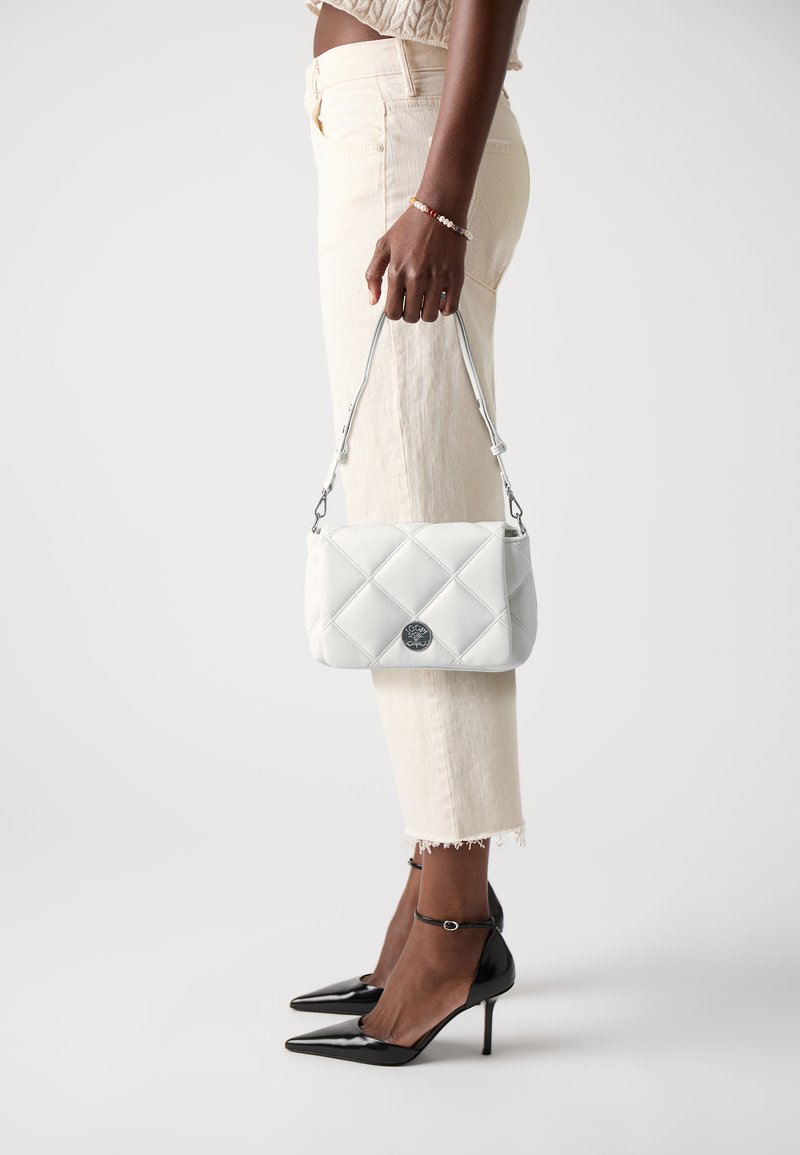 Borsa a tracolla bianca trapuntata con logo argentato, dotata di una maniglia corta, sostenuta di lato. Il modello indossa pantaloni chiari e tacchi alti neri.