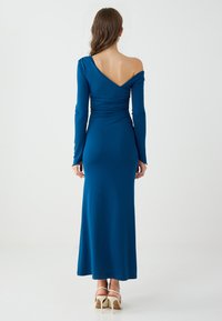 Robe à manches longues, asymétrique, décolletée sur l'épaule en bleu sarcelle. Présente un ruché dans le dos et une silhouette cintrée. Tissu lisse et longueur au sol.