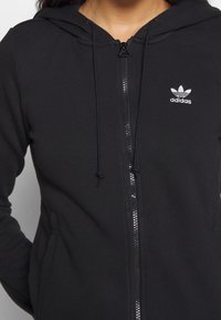 Svart zip-up hoodie i mjukt tyg, med dragsko på huvan och en liten vit tryckt logotyp på vänster bröst.