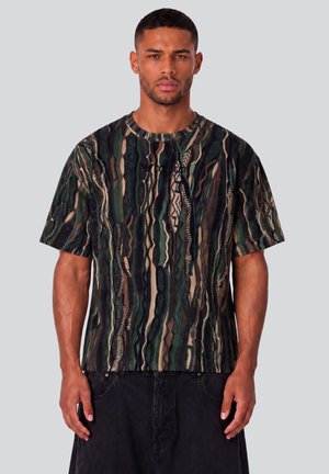 Hombre con camisa de manga corta de varios colores y pantalones negros, de pie frente a un fondo claro y liso.