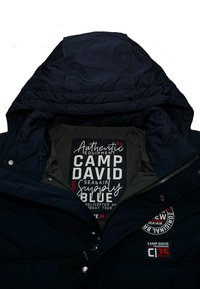 Chaqueta acolchada de color azul marino con capucha desmontable, textura acolchada y parches con detalles de marca en un forro gris.