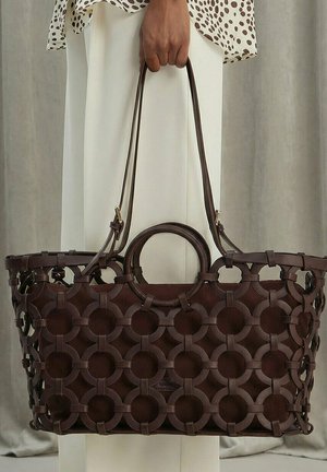 Mano sosteniendo un bolso tote grande de cuero marrón oscuro con patrón de cortes circulares y doble asa, sobre pantalones blancos y parte superior estampada.