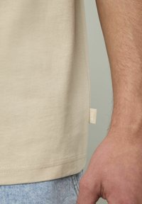 T-shirt en coton beige à coupe droite, avec une étiquette sur la couture latérale et des manches courtes, associé à un jean en denim bleu clair. Texture lisse.