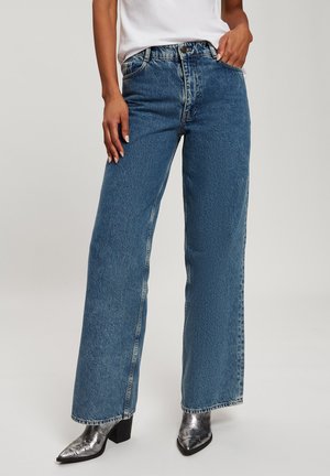 Wide-leg blue denim jeans with a faded texture, high waist, front pockets, and silver hardware, paired with metallic heeled boots.