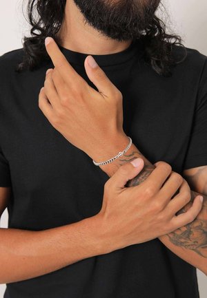 Silver chain bracelet with a round charm, worn on a wrist with visible tattoos, against a plain black t-shirt background.