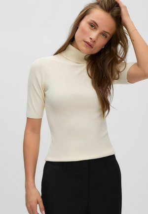Femme aux longs cheveux bruns portant un col roulé court côtelé crème et un pantalon noir, posant avec une main dans les cheveux sur un fond uni.