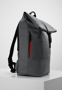 Szary plecak z tkaniny z zamknięciem typu roll-top, czarnymi paskami, czerwoną kieszenią na zamek błyskawiczny i eleganckim designem. Gładka faktura z minimalnym brandingiem.