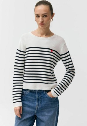 Kobieta z włosami związanymi do tyłu, nosząca złote kolczyki koła, biało-czarny sweter w paski z małym czerwonym sercem oraz niebieskie jeansy, z ręką w kieszeni.