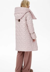 Jasnoróżowy pikowany płaszcz z dużym kapturem, z teksturowanym wzorem i skórzaną naszywką z logo, zestawiony z dopasowanymi, luźnymi spodniami.