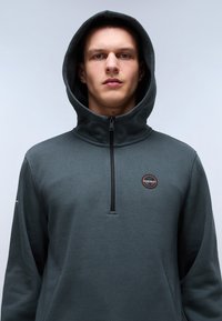 Mørkegrøn pullover hoodie med høj krave, lynlås lukning og et cirkulært logo mærke. Blødt stof og tekstureret overflade. Har en frontlomme.