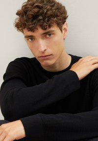 Pullover in maglia nera con texture e scollo rotondo. Le mani del modello sono appoggiate sulle braccia, evidenziando le maniche lunghe e il design aderente. Sfondo chiaro.
