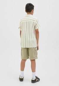 Kortsleeve, krämfärgad randig skjorta i lätt tyg, kombinerad med khaki cargo-shorts. Bruna sneakers och vita strumpor fulländar outfiten.