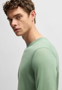 Hellgrüner Strickpullover mit Rundhalsausschnitt, der eine schlanke Passform, eine glatte Textur und sichtbare Nähte entlang der Schultern aufweist.