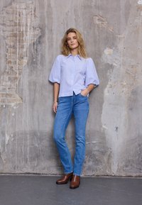 Gestreepte lichtblauwe blouse met pofmouwen, gecombineerd met strakke blauwe spijkerbroek en bruine laarzen met hak, tegen een getextureerde betonnen muur.