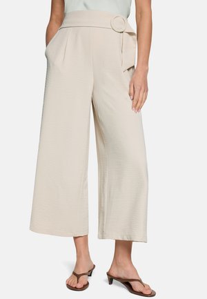 Femme portant un pantalon beige taille haute à jambes larges avec une boucle de ceinture circulaire et des sandales à talons marron, debout devant un fond uni.