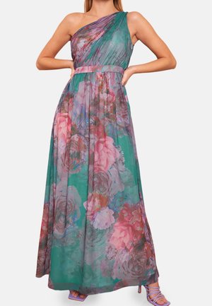 Femme portant une robe longue asymétrique à une épaule, avec des motifs floraux roses et violets sur un fond vert, assortie à des talons à lanières violets.