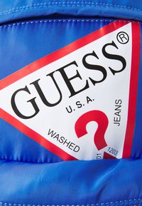 Veste bleue arborant un logo triangulaire avec "GUESS U.S.A." et un point d'interrogation rouge, sur un fond lisse et brillant.