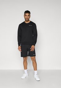 Sweatshirt noir avec un col rond, associé à un short noir. Les deux présentent un petit logo et une texture lisse. Des sneakers blanches complètent le look.