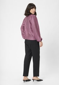 Helle, glänzende lila Bluse mit gepufften Ärmeln, kombiniert mit schwarzen Hosen und spitzen schwarzen Mules, von hinten betrachtet.