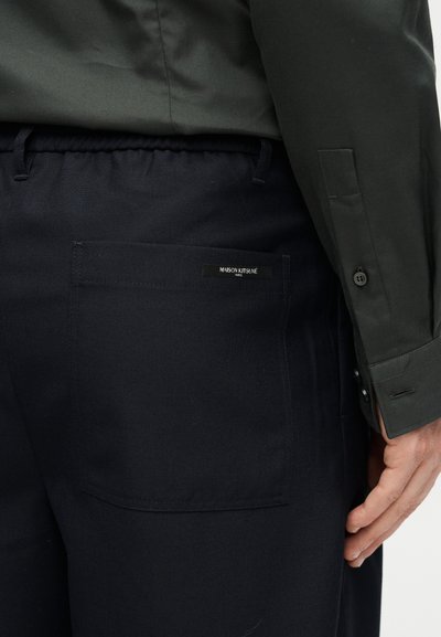 Κοντινό πλάνο ενός ατόμου που φοράει μαύρο παντελόνι με πίσω τσέπη που φέρει την ετικέτα "Maison Kitsuné", σε συνδυασμό με σκούρο πράσινο μακρυμάνικο πουκάμισο.