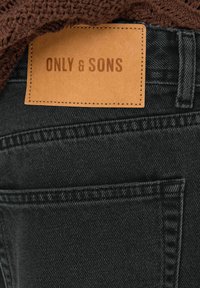 Crne traperice od denima s smešnim kožnim oznakom "ONLY & SONS." Imaju otkrivenu šavovu obradu i teksturirani smeđi pleteni sloj iznad.
