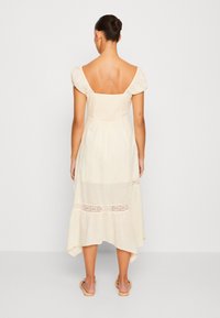 Woman with hair in a bun wearing a cream, square-neck, short-sleeve midi dress with lace panels, standing barefoot against a plain background.