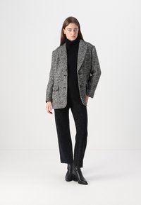 Blazer en pied de poule texturé noir et blanc, superposé à un col roulé noir ajusté, associé à un pantalon noir avec un subtil éclat.