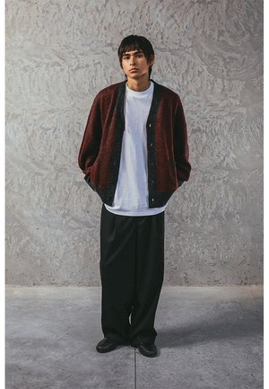 Dunkelroter Wollcardigan über einem weißen T-Shirt, kombiniert mit lockeren schwarzen Hosen. Mit Knöpfen, Seitentaschen und einer lässigen Passform.