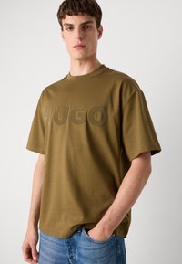 Olive roheline puuvillane T-särk, millel on mugav lõige, esiküljel on suur, muljetavaldav "HUGO" logo. Lühikesed varrukad ja ümar kaelus.